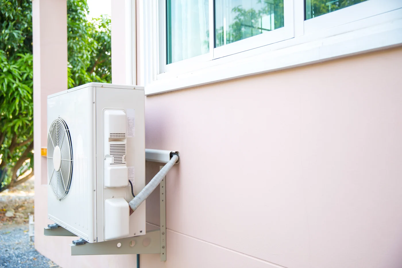 A white AC unit sits outside a house with light pink walls. There are some plants around the house.