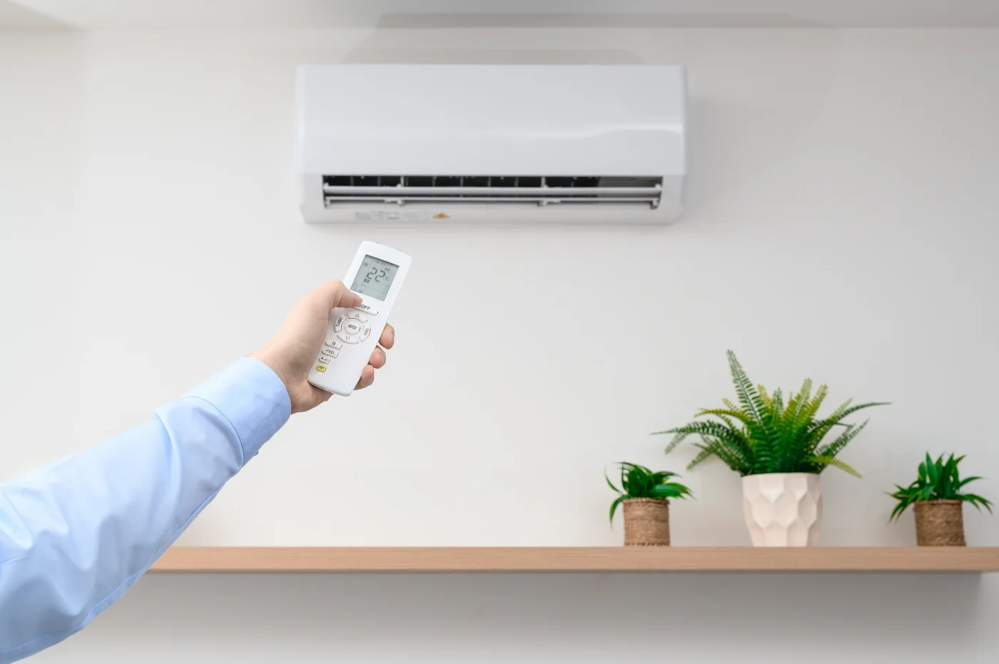 A person's hand holds a remote control pointed at a wall-mounted AC unit, with decorative green plants arranged neatly on a shelf below.