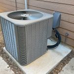 A central air conditioning unit is installed on a concrete pad beside a house with brown siding. The unit’s fan is visible on top, and insulated refrigerant lines connect it to the home through an exterior wall. The surrounding area is neatly landscaped with gravel and concrete pathways, emphasizing a clean and well-maintained installation