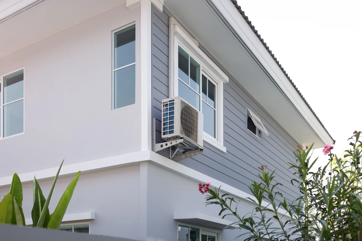 The exterior of a modern two-story home with light gray siding and white trim. Mounted just below an upper-story window is a wall-mounted outdoor AC unit, part of a split air conditioning system. In the foreground, green plants and pink flowers partially frame the view, adding a touch of natural color to the clean, contemporary house design