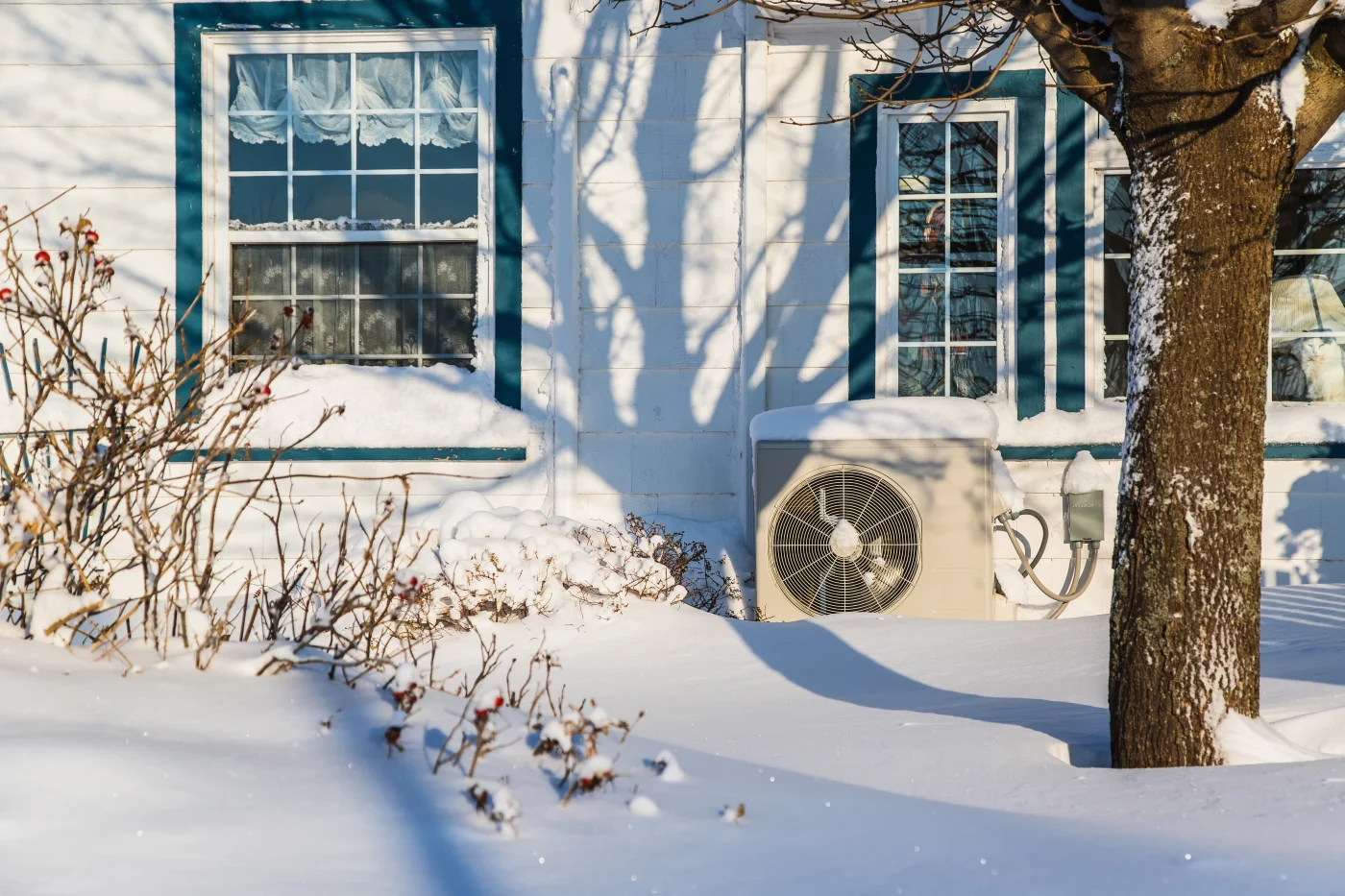 An outdoor heat pump unit sits beside a snow-covered home, framed by frosty windows and bare winter branches. The scene highlights reliable year-round heating performance, even in the harsh Minnesota cold
