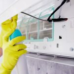 A tech wearing bright yellow protective gloves is cleaning the interior of a wall-mounted air conditioning unit. The front panel of the AC is open, revealing the filter and internal components. The technician is holding a spray bottle aimed at the filter, suggesting maintenance or sanitization