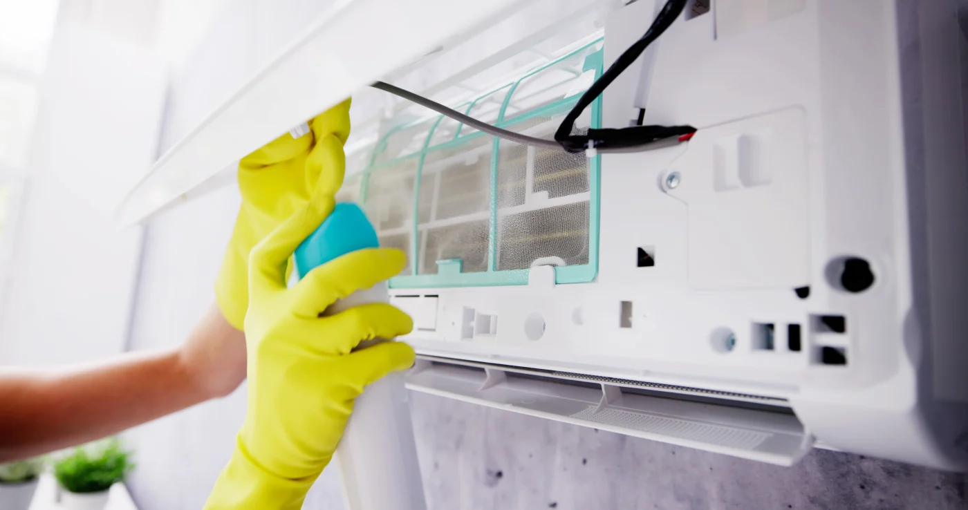 A tech wearing bright yellow protective gloves is cleaning the interior of a wall-mounted air conditioning unit. The front panel of the AC is open, revealing the filter and internal components. The technician is holding a spray bottle aimed at the filter, suggesting maintenance or sanitization