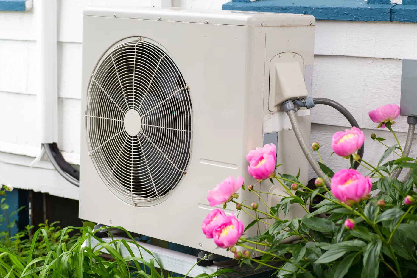 A beige outdoor heat pump unit sits beside a house, surrounded by blooming pink flowers and green plants, highlighting its role in efficient home heating and cooling