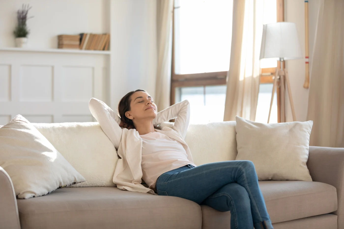 A woman relaxes on a beige sofa with her hands behind her head and eyes closed, enjoying the comfort of a cozy, sunlit living room.