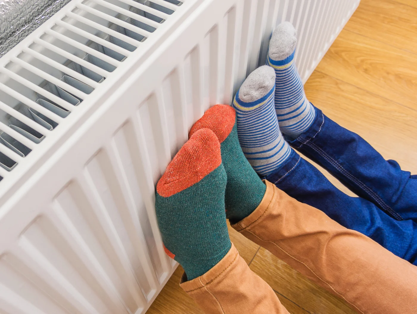 Two pairs of sock-covered feet rest against a white radiator, soaking in the warmth. One person wears green and red socks with tan pants, while the other wears blue striped socks with dark jeans. The cozy indoor scene emphasizes the comfort and reliability of a well-maintained heating system.