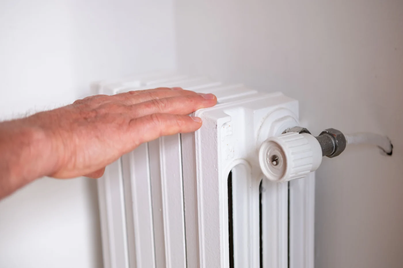 A close-up of a person's hand hovering just above a white, cast-iron radiator. The radiator's valve and connecting pipe are visible, and the scene suggests someone checking the radiator's warmth. The white wall and radiator create a clean, simple backdrop, emphasizing the action of testing the heat.