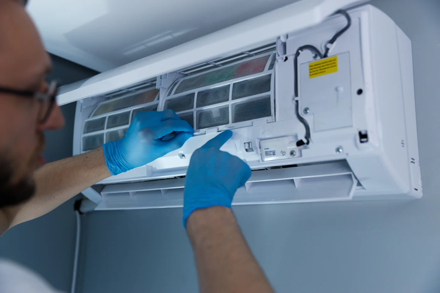 A technician wearing blue gloves works on the interior filter of a wall-mounted air conditioning unit, carefully adjusting components during a repair or maintenance service.