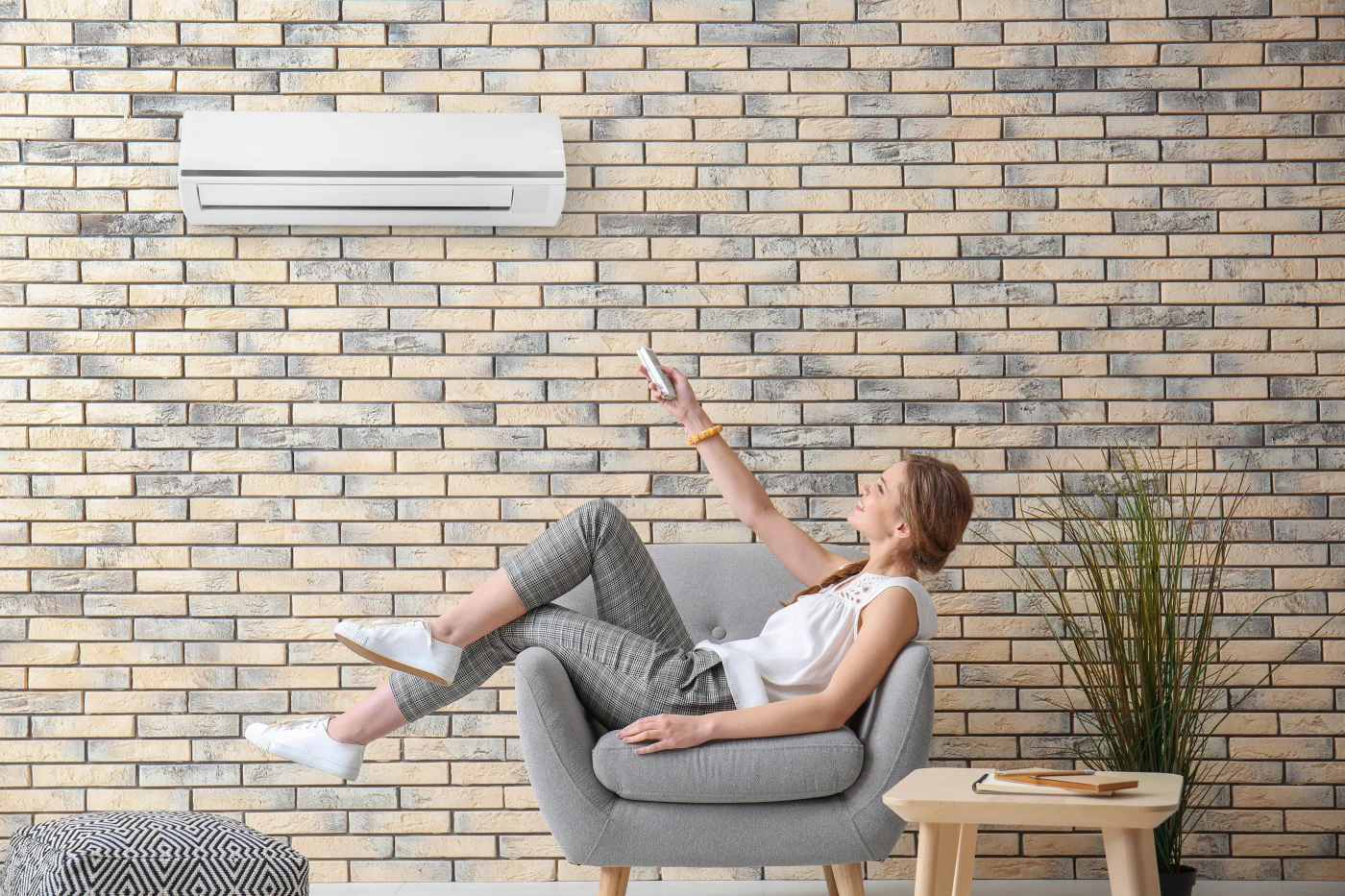 A woman relaxes in a modern living room, pointing a remote control toward a wall-mounted air conditioning unit. She’s seated comfortably in a gray armchair against a brick accent wall, enjoying the cool, even airflow