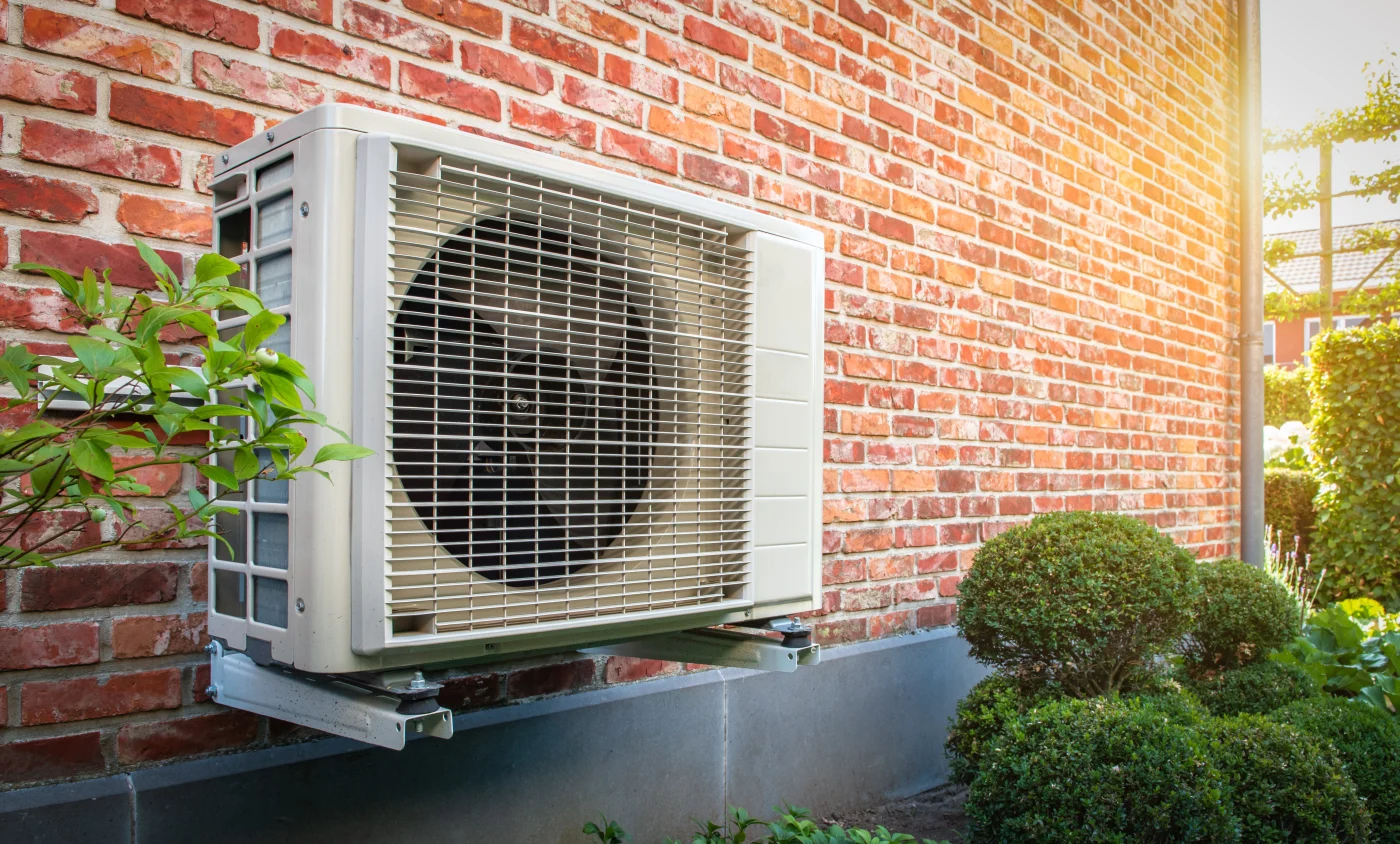 A beige outdoor heat pump unit is mounted on a brick wall above trimmed bushes, with greenery and sunlight surrounding the system.
