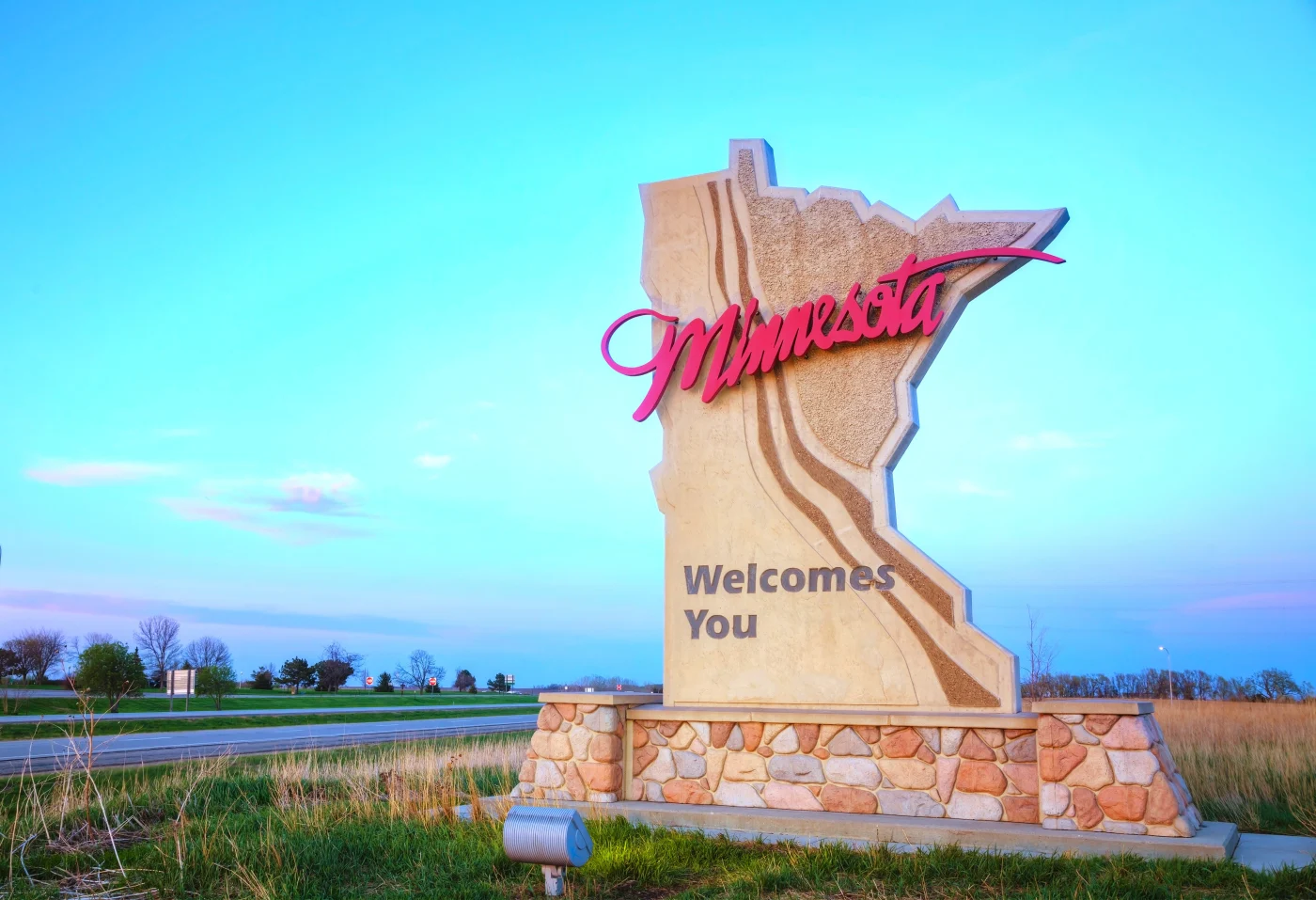 A large roadside monument sign in the shape of Minnesota greets travelers with the words “Minnesota Welcomes You” in bold letters. The sign features a textured stone base and a stylized script for “Minnesota” in bright red. Against a clear blue sky and open landscape, it conveys a warm welcome to the state