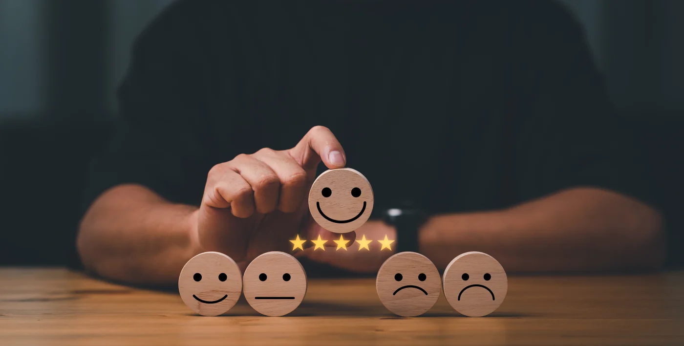 A hand selects a wooden disc with a smiling face on it from a row of expression-faced discs. The smiley disc is highlighted with five gold stars below it, representing a top-rated or five-star review. The dark background keeps the focus on the wooden discs and the gesture of choosing the happy, satisfied face.