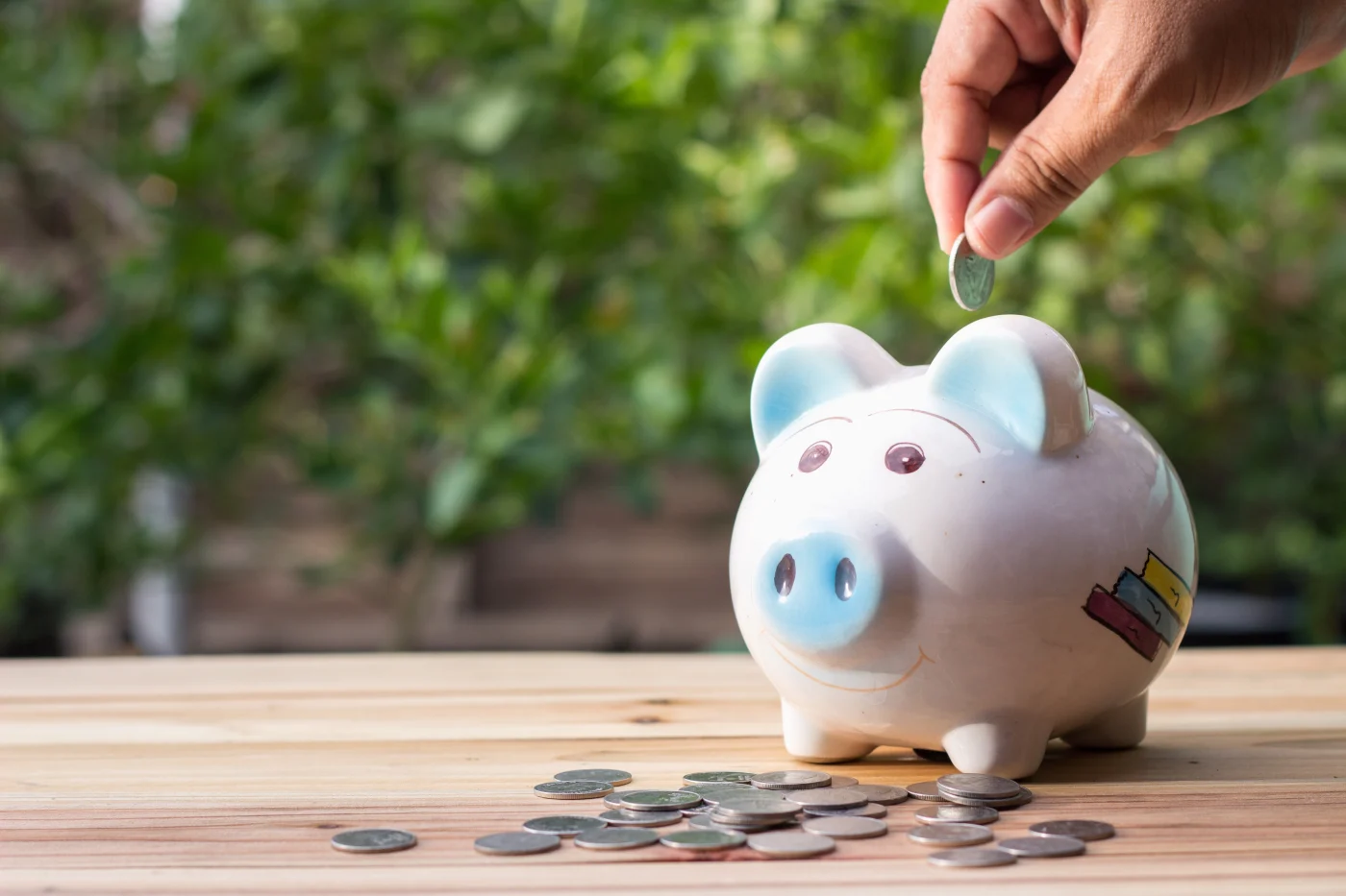A close-up of a hand placing a coin into a white piggy bank with light blue accents on its ears and snout. The piggy bank sits on a wooden surface, surrounded by a small pile of coins. The background is blurred greenery, giving the photo a fresh, outdoor feel. This image conveys themes of saving money on AC replacement