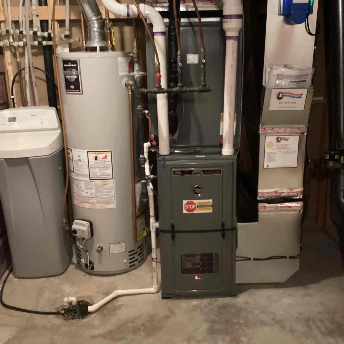 Residential furnace system in a basement mechanical room, showing a gas furnace, water heater, connected PVC venting, and drainage piping.