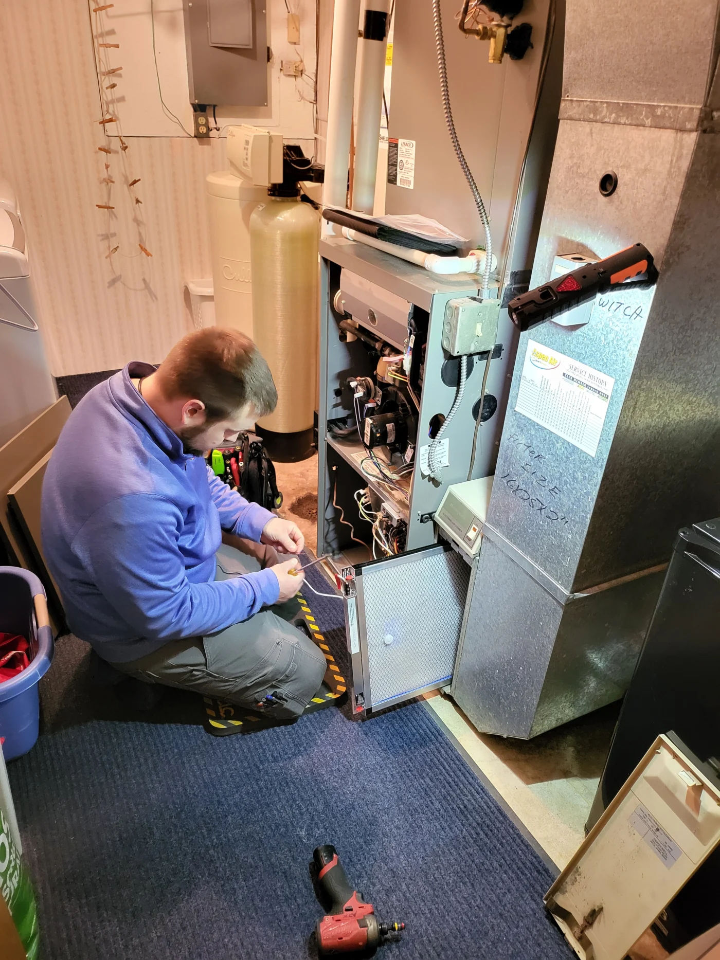 Technician in blue uniform kneeling on a basement floor while performing detailed furnace service on an open heating unit, with tools and wiring.