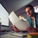 Focused man in glasses sitting at a desk in a dimly lit home office, reviewing paperwork in front of a large computer monitor.