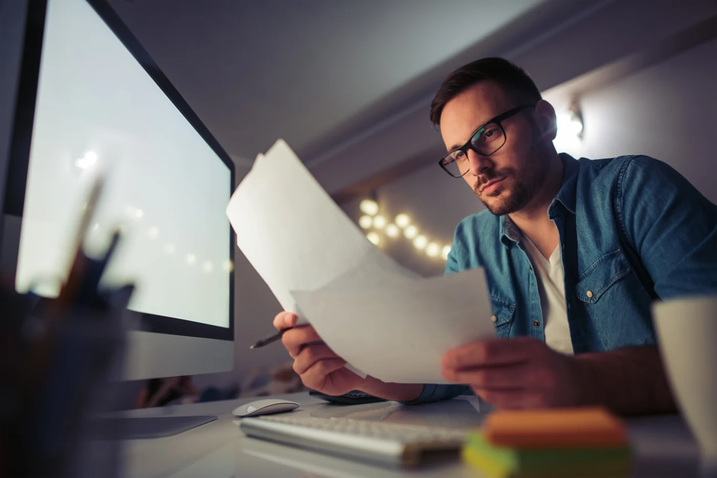 Focused man in glasses sitting at a desk in a dimly lit home office, reviewing paperwork in front of a large computer monitor.