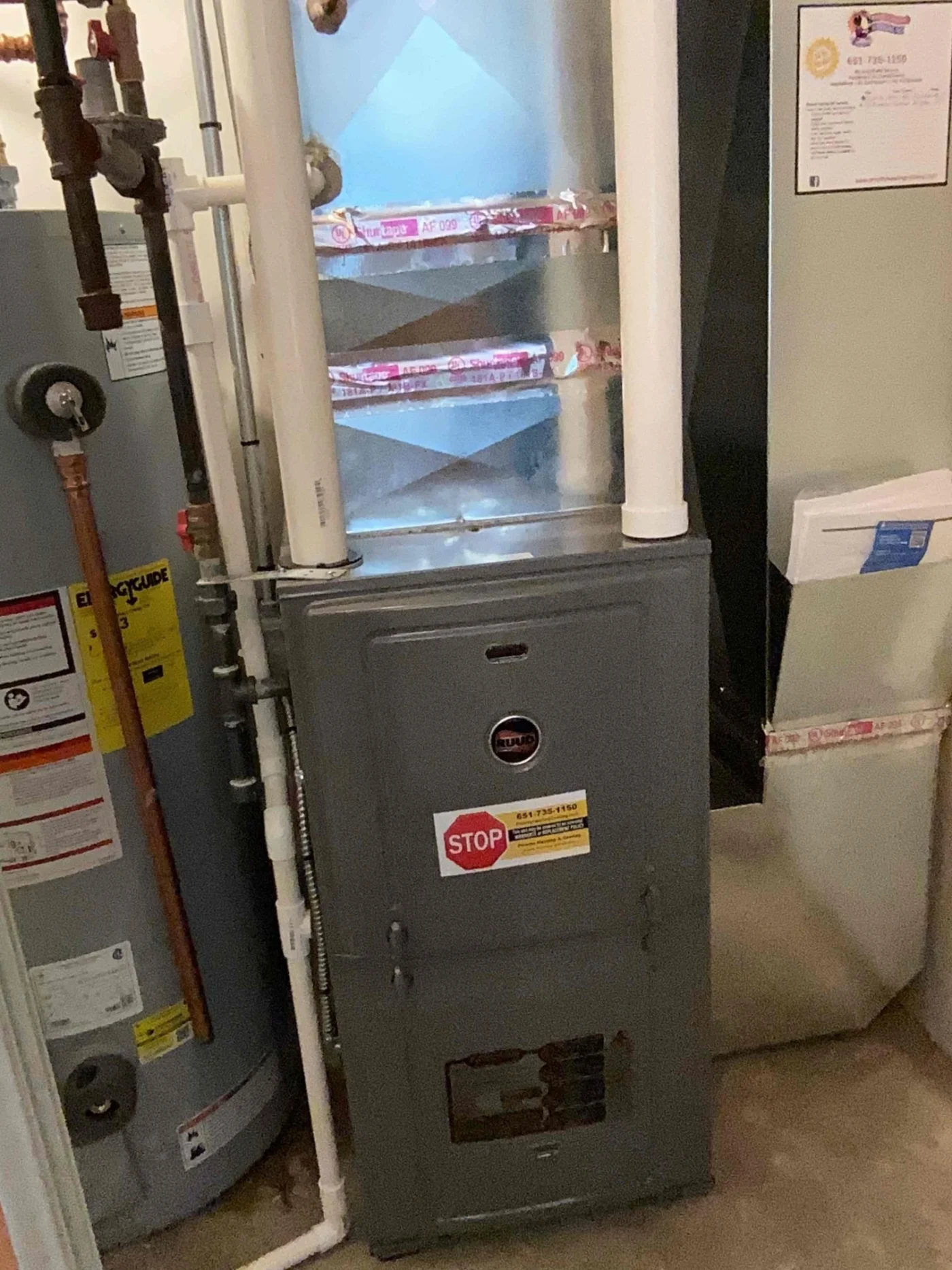 A gray furnace unit beside a water heater and ductwork in a utility room with exposed pipes and service labels.