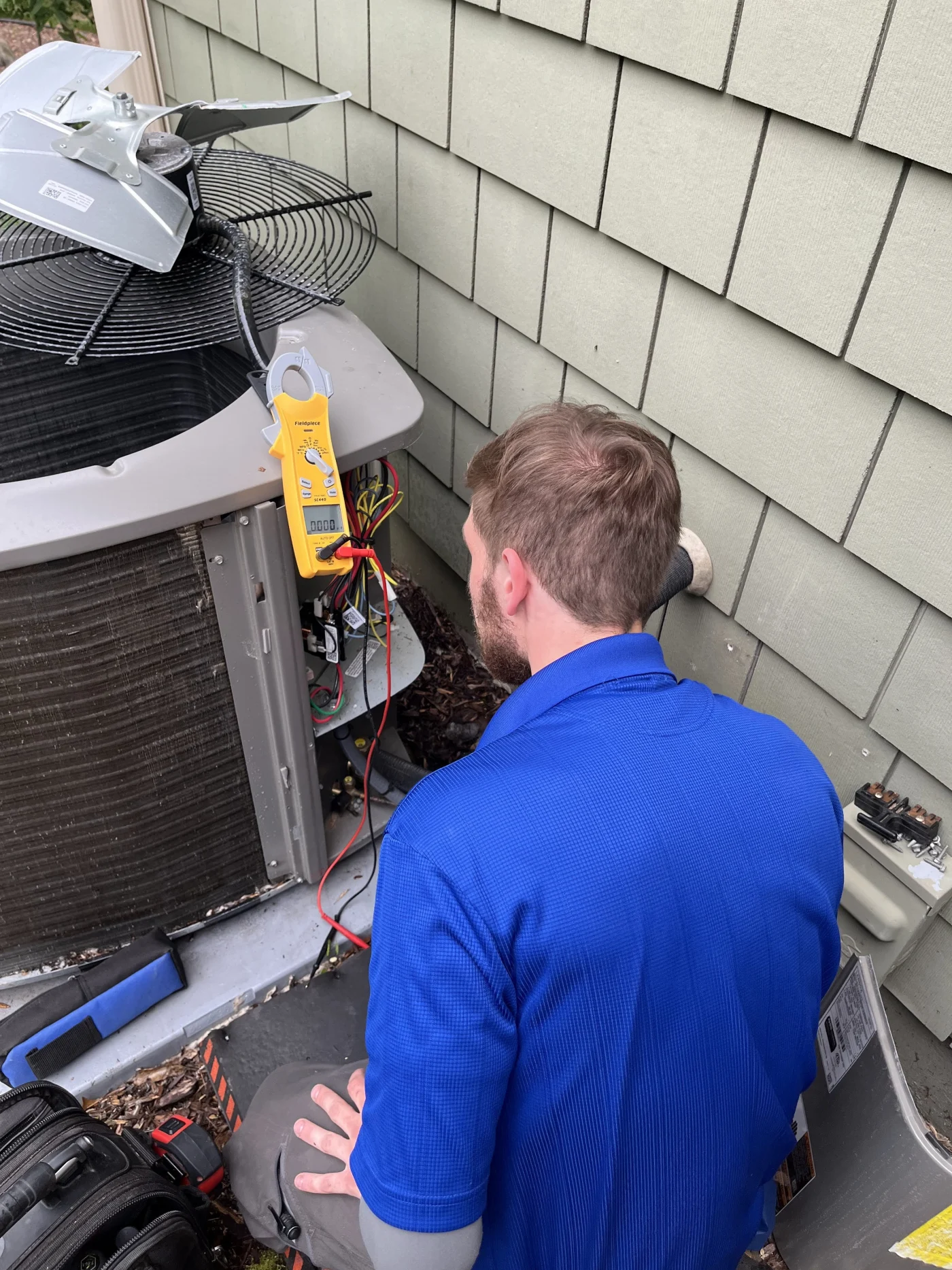 Expert AC Service | Maplewood, MN | Priority Heating and Cooling An HVAC technician in a blue shirt is servicing an outdoor air conditioner condenser, using a clamp meter and tools beside a home's exterior wall.
