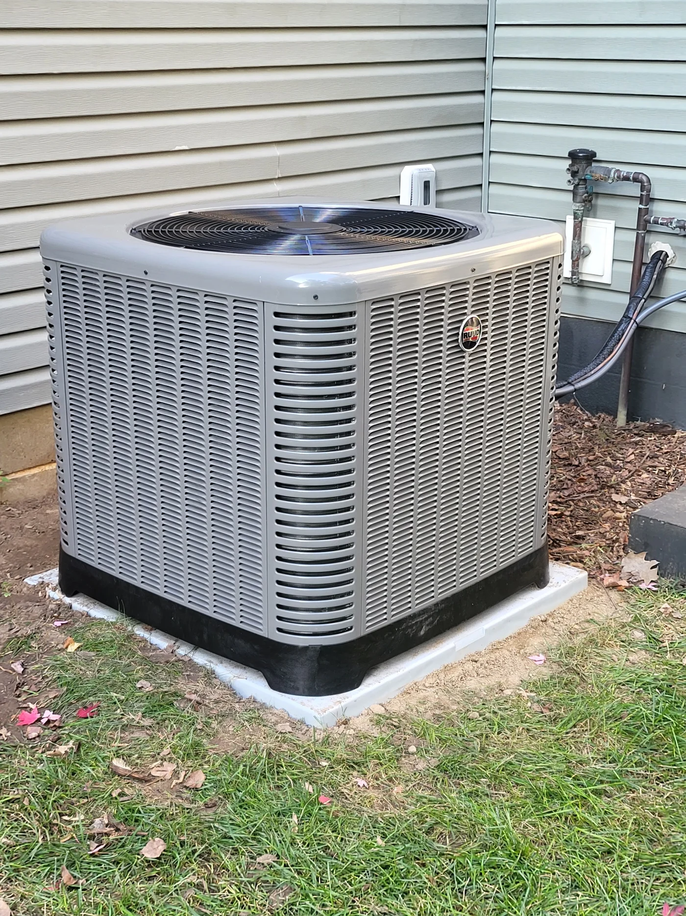 Ruud outdoor air conditioning unit installed beside a home with light siding, utility lines, mulch, and grass nearby.