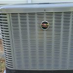 Close-up of a Ruud outdoor air conditioning unit installed beside a home with light siding and a downspout.