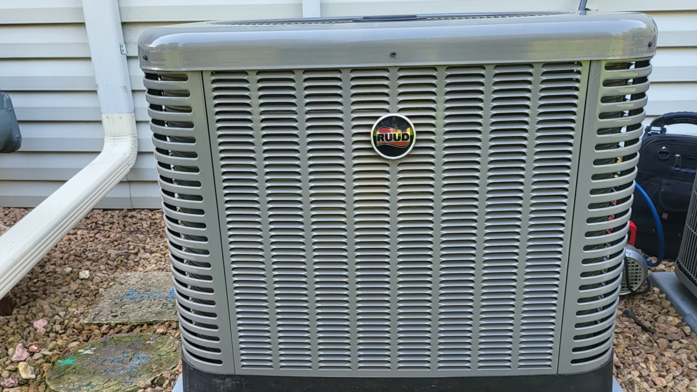 Close-up of a Ruud outdoor air conditioning unit installed beside a home with light siding and a downspout.