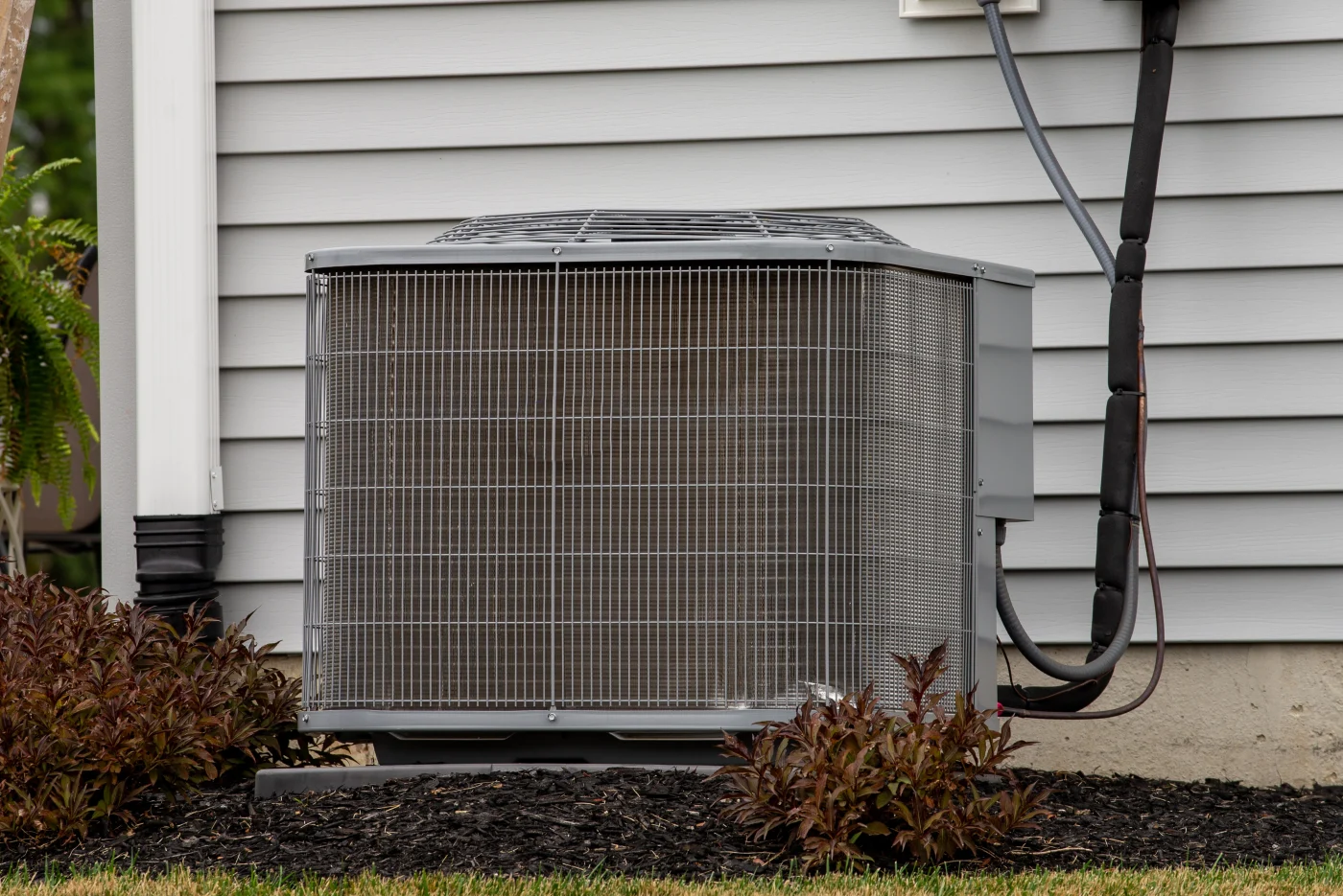 An outdoor central air conditioning unit is installed next to a home’s exterior wall, with visible wiring, mulch landscaping, and small shrubs.