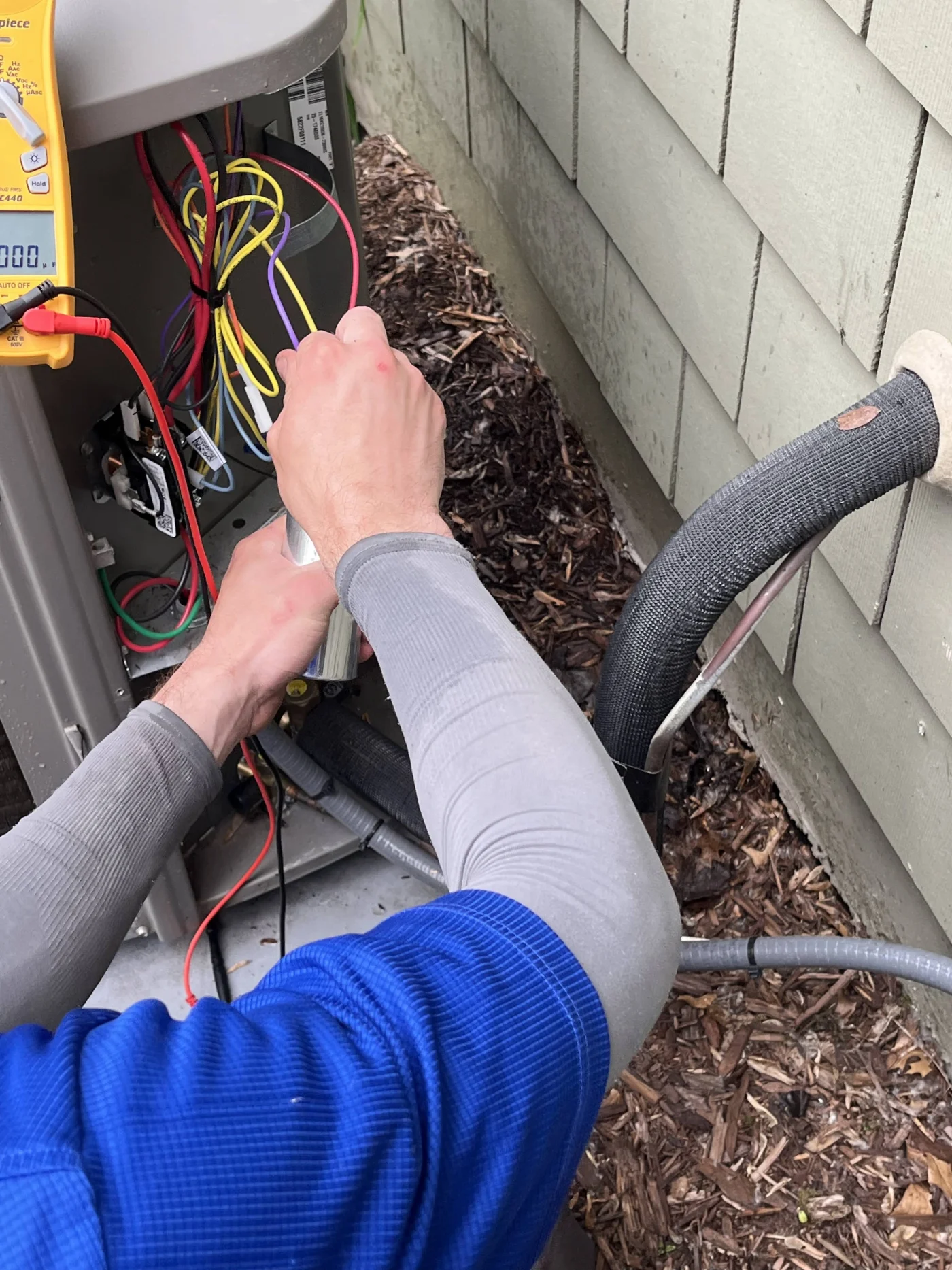 Master AC Service | Maplewood, MN | Priority Heating and Cooling A close-up of an HVAC technician replacing or wiring a capacitor inside an outdoor air conditioner condenser while testing components with a digital clamp.